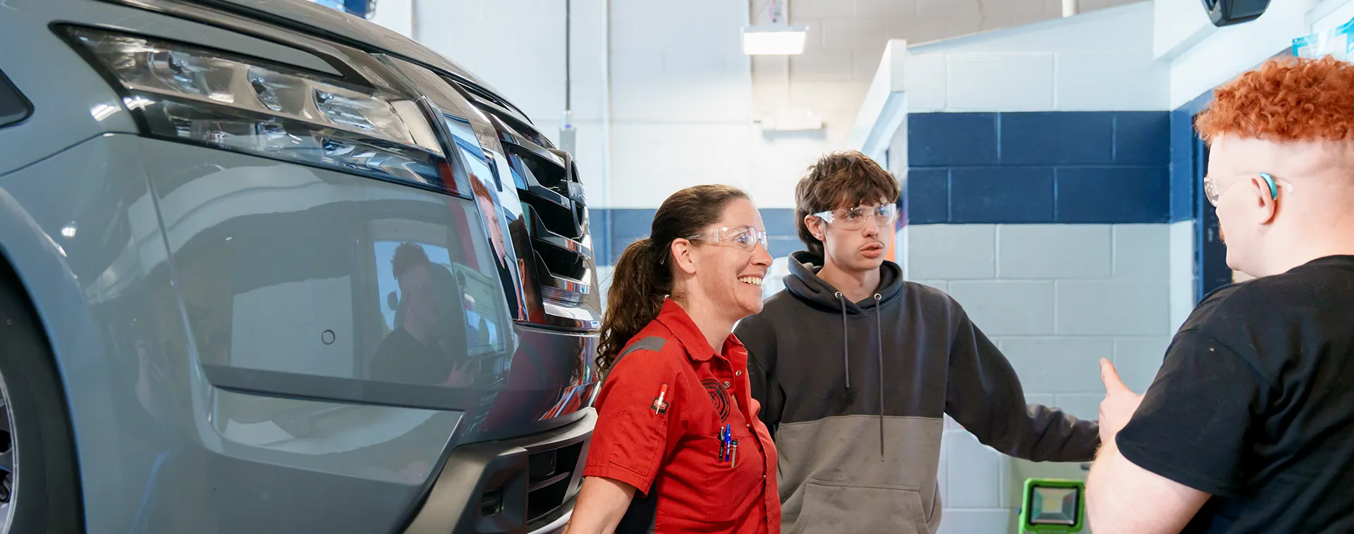Instructor and two students wearing safety glasses stand beside a raised vehicle in an automotive lab.