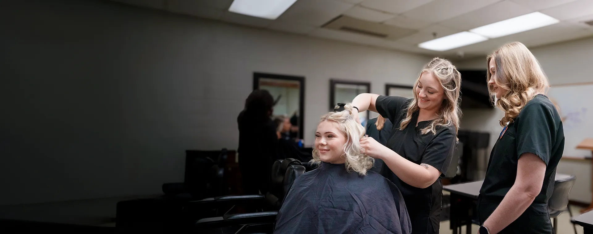 Two students style a seated client&rsquo;s hair in a classroom salon with mirrors and overhead lighting.