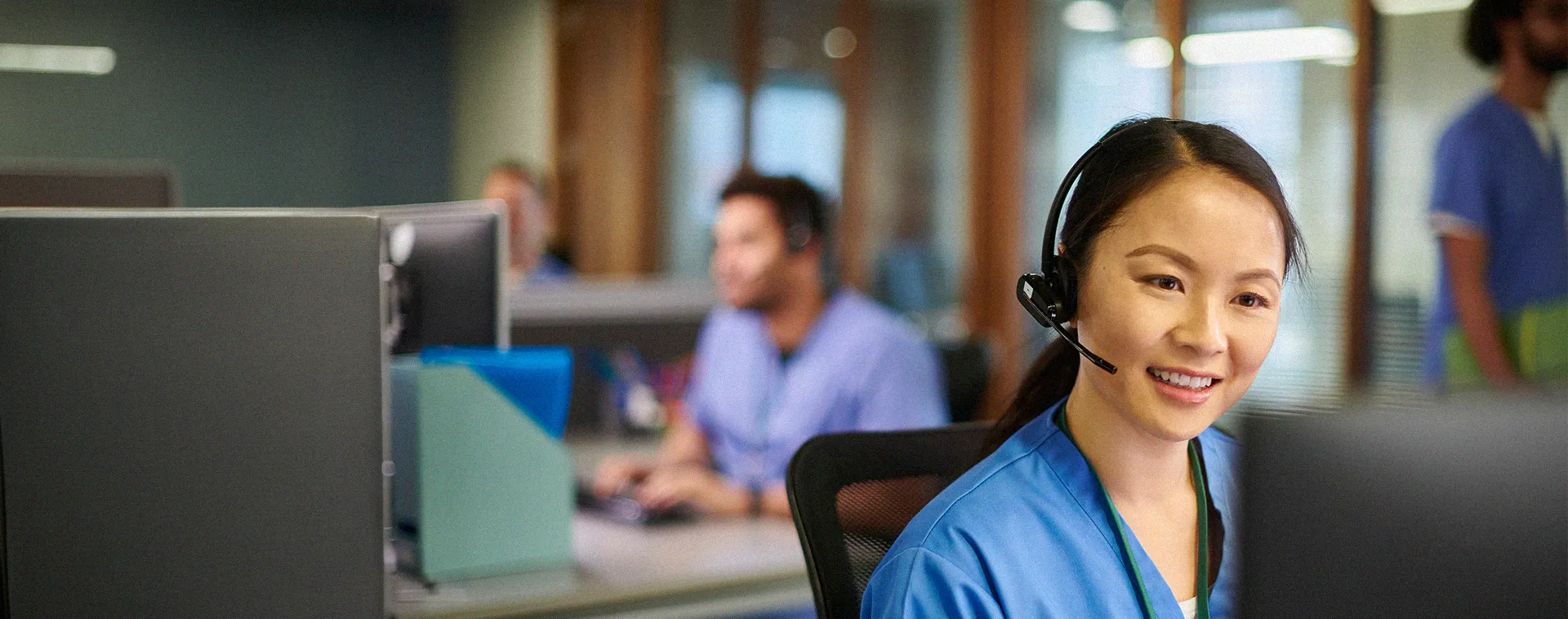Person wearing a headset using a computer in an office, with coworkers and desks blurred in the background.
