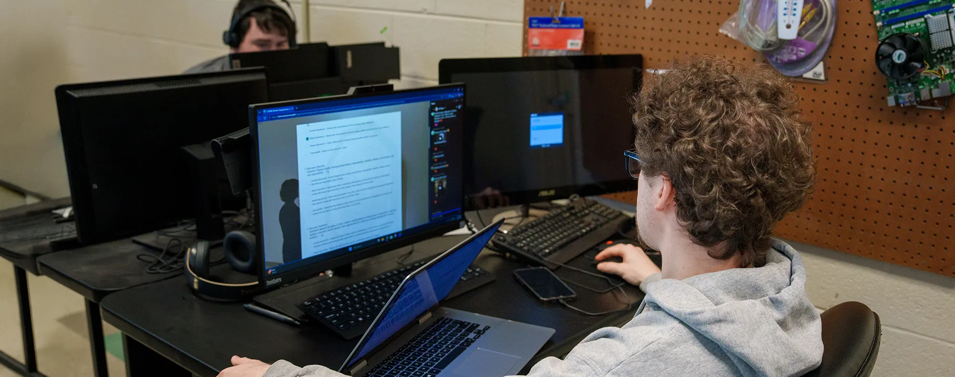 Student at a workstation using dual monitors and a laptop, with headphones, phone, and multiple desktop computers on a desk.