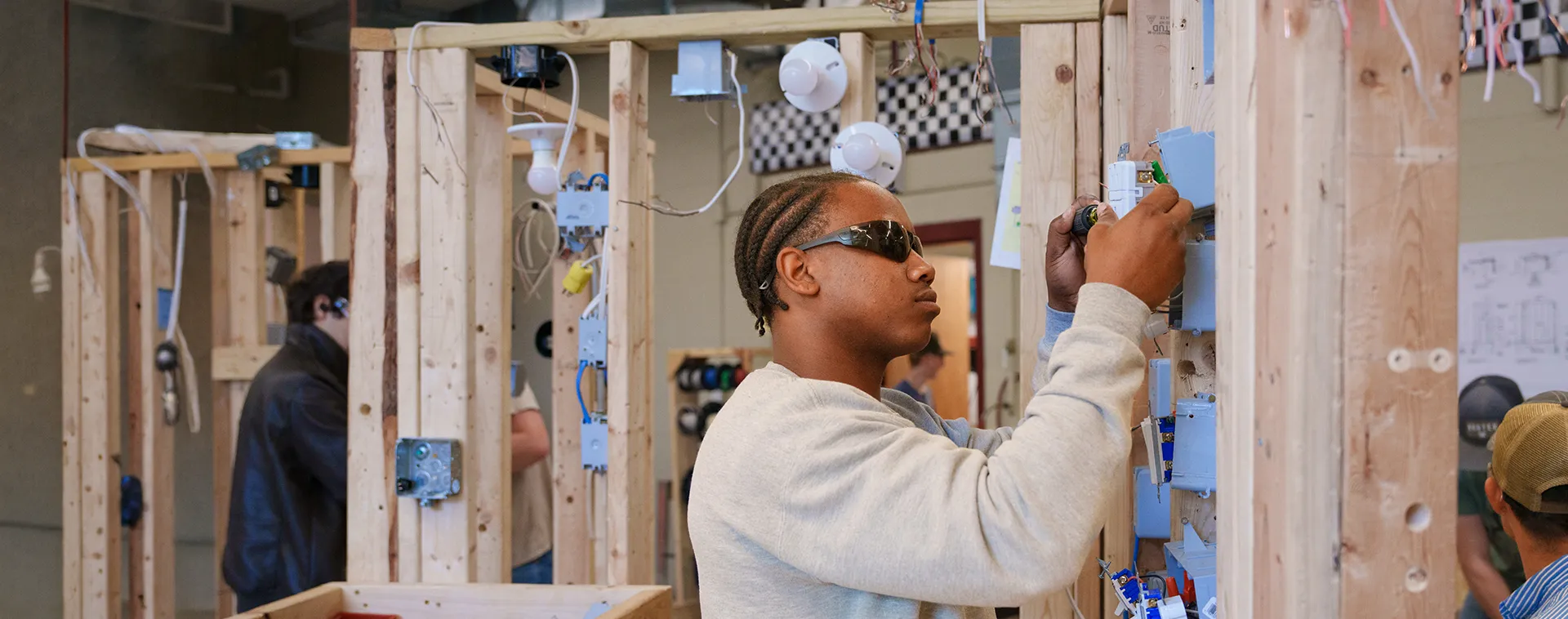 Student wearing safety glasses installs wiring in a wooden wall training frame with outlets, switches, and cables.