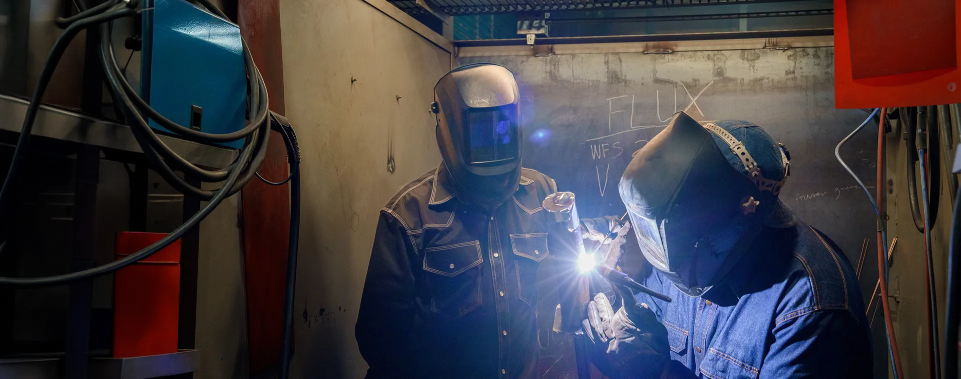 Two people wearing welding helmets work together at a station, with a bright welding arc and equipment in a workshop.