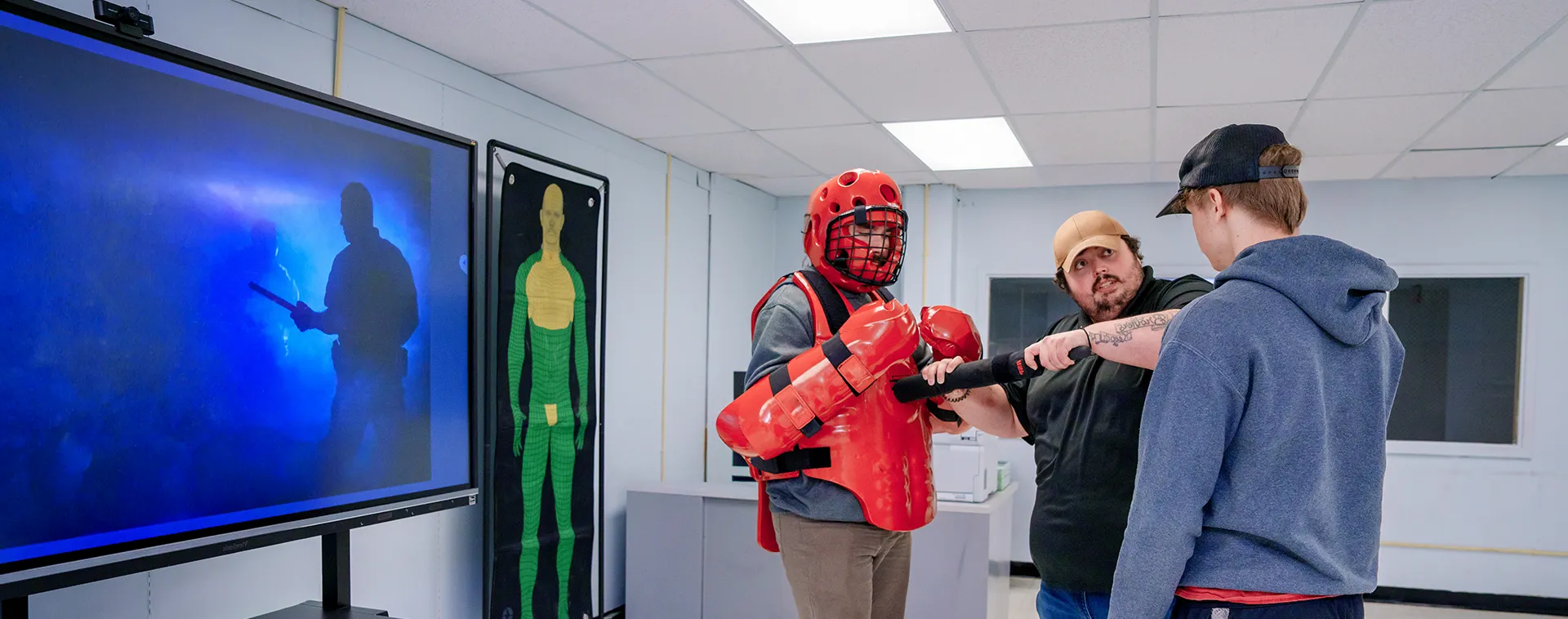 Instructor guides two students using batons and protective gear in a classroom with a screen and target graphic on the wall.
