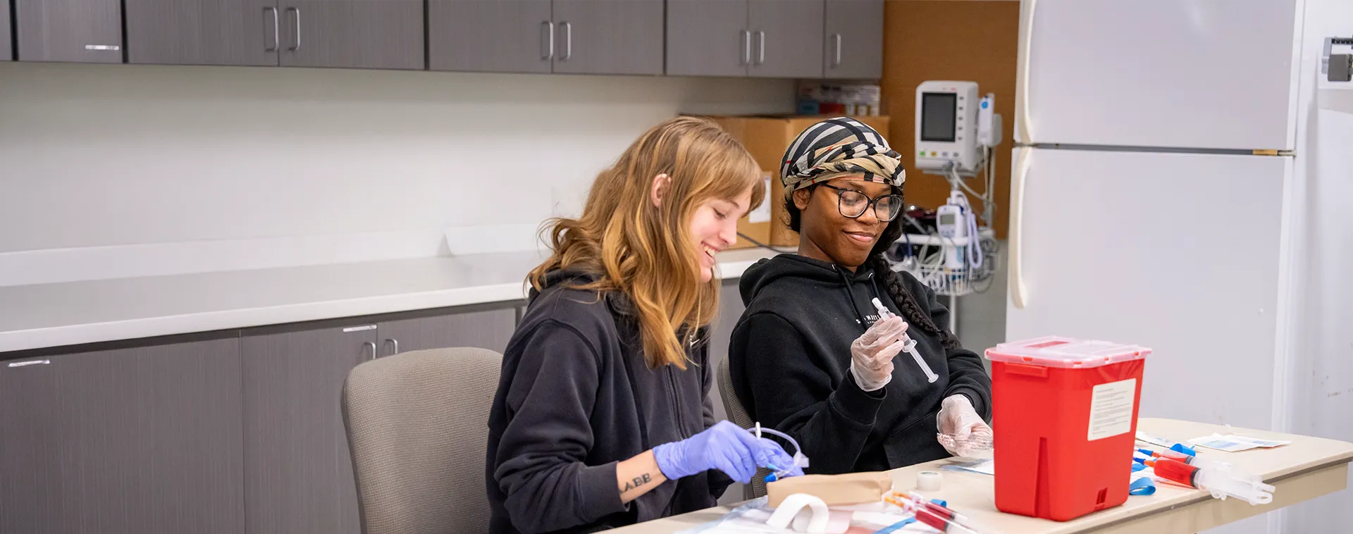 Two students seated at a table practice with syringes and medical supplies, wearing gloves, with cabinets and a red sharps container nearby.