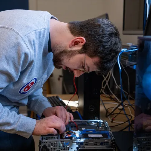 Person wearing safety glasses connects wires inside an open desktop computer with tools and cables on a workbench.
