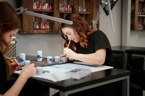 Student files nails at a workstation with small jars of nail products and tools on the table and shelves behind.