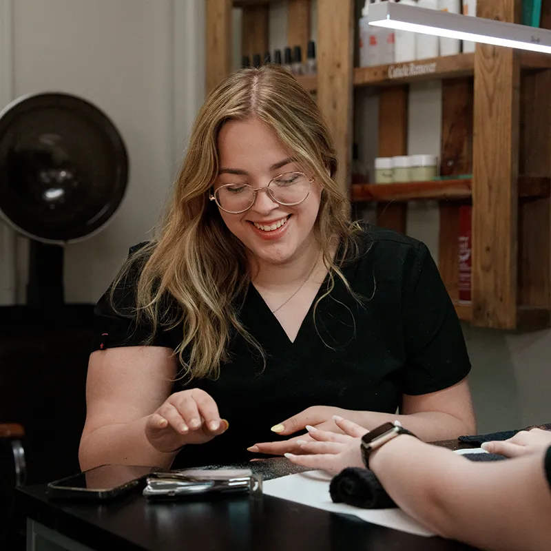 Person in black top applying manicure to another hand at a table, with a smartphone, tools, towel, and wooden shelves in background