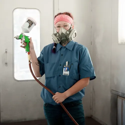 Student wearing a respirator holds a paint spray gun with hose inside a vehicle paint booth