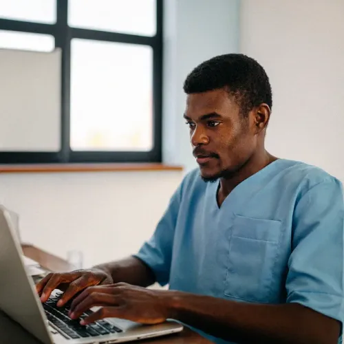 Person in blue scrubs typing on a laptop at a wooden desk near a window with black grid panes.