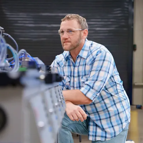 Person wearing safety glasses kneels beside a machine with tubes and components in a lab or workshop setting.