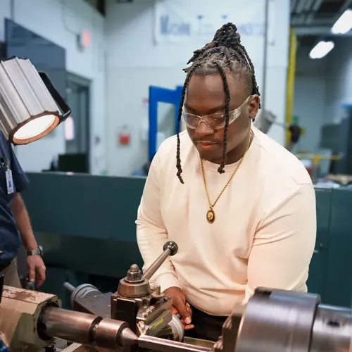 Student wearing safety glasses operates a metal lathe, adjusting controls under a task light in a workshop.