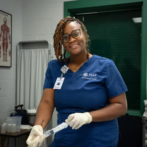 Person in blue scrubs with gloves holds a syringe in a clinical room with a privacy curtain, anatomy poster, and medical equipment visible.