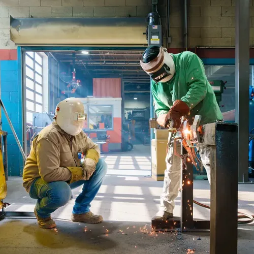 Two people wearing welding helmets cut or weld metal on a workbench, with sparks and tools visible in a shop setting.