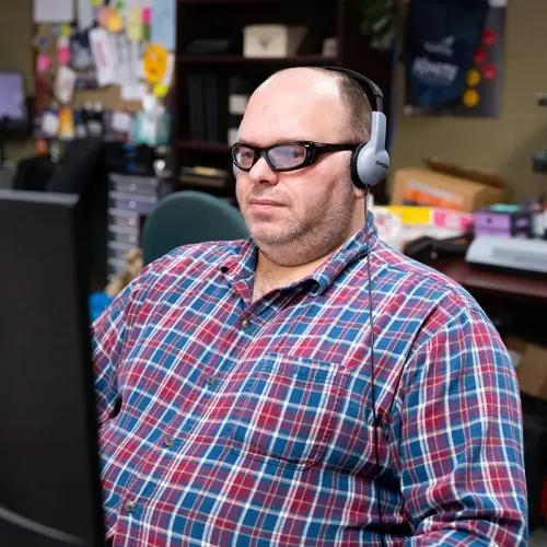 Person wearing headphones and glasses, seated at computer workstation in office with shelves and supplies visible