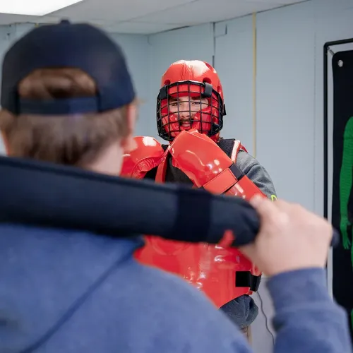 Student in red protective gear faces another holding a training baton in a classroom with a wall-mounted target display.