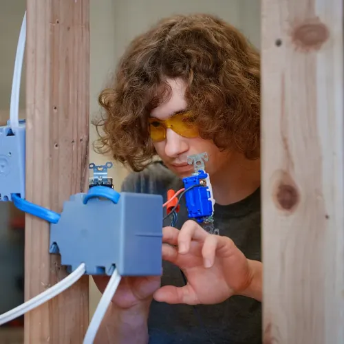 Student wearing yellow safety glasses connects wiring to an electrical box in a wood-framed training wall.