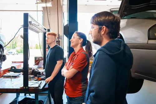 Three students observe a computer diagnostic setup near a vehicle lift in an automotive training shop
