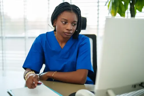 Person in blue scrubs sitting at a desk with a desktop computer, clipboard, and plant near a window with blinds.