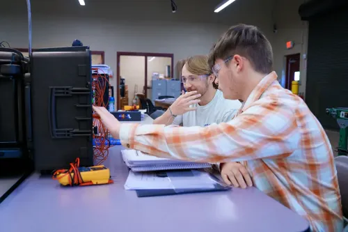 Two students wear safety glasses while working with wiring and tools at a lab bench with notebooks and equipment.