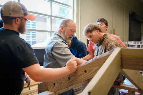 Instructor and students assemble a wooden structure frame in a workshop with tools and lumber.