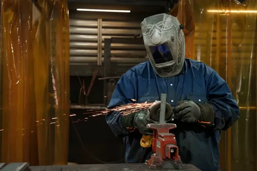 Person in protective gear uses a grinder on metal, with sparks flying in a workshop enclosed by safety curtains.