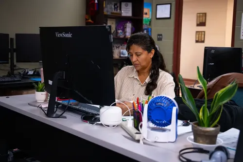 Person working at desk with computer, small fan, plant, and office supplies; shelves and doorway behind.
