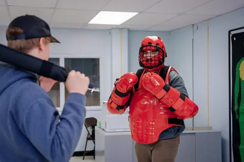 Instructor gestures while two students stand nearby, one wearing red padded protective equipment during a training exercise.