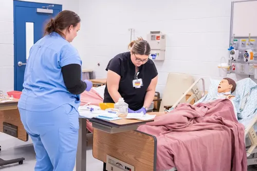 Two learners in scrubs stand beside a hospital bed, preparing supplies while practicing on a mannequin in a clinical classroom.