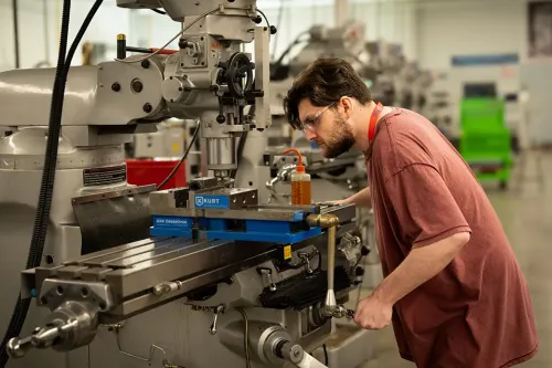 Student wearing safety glasses operates a vertical milling machine with metal components in a workshop.